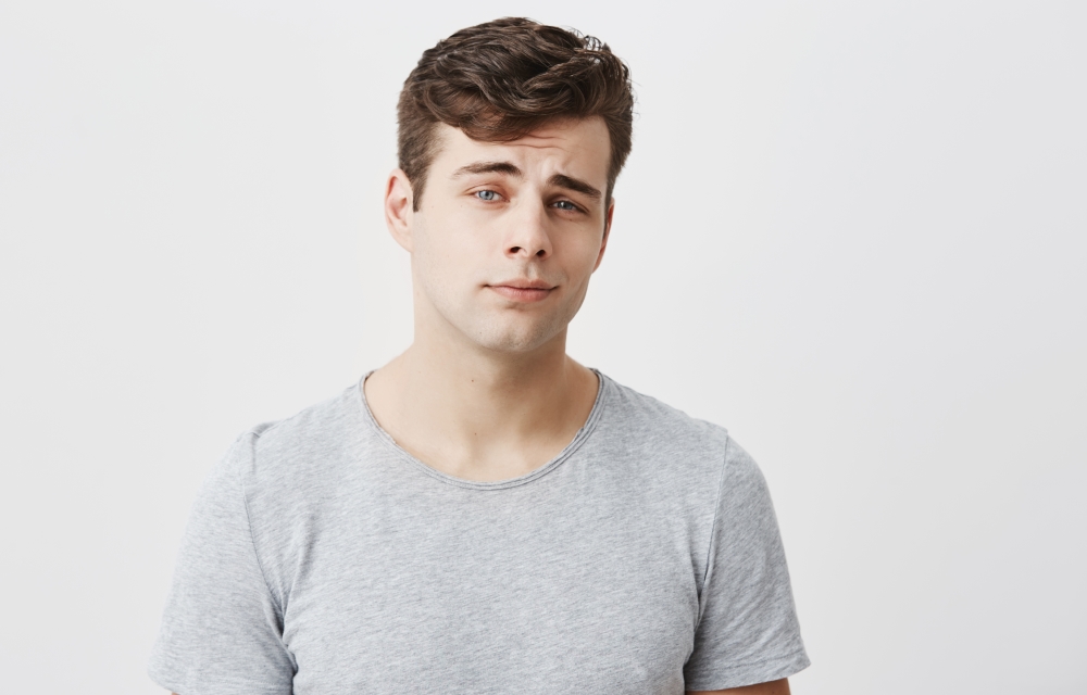 Good-looking stylish handsome male posing against gray studio wall. Attractive caucasian guy dressed casually with dark hair, looks at camera with his blue eyes, has thoughtful face expression.